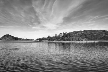 Norveç 'te açık denizin önündeki fiyortta kayalar ve ağaçlarla dolu bir ada. Su güneşte parlıyor. Kuzeyden manzara.