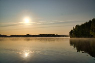 İsveç gölünün üzerinde gün doğumu. Sahildeki ağaçların önünde ayna gibi pürüzsüz su var. İskandinavya 'dan doğa çekimi