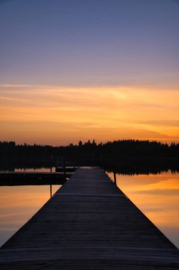 İsveç gölünde gün batımında ahşap iskelede. Günbatımının renkleri suya yansıyor. İskandinav kırsalında bir banyo gölü