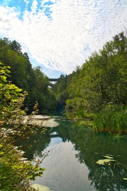 Yoğun bitki örtüsüyle kaplı sakin bir nehir dramatik bulutlarla gökyüzünü yansıtır. Arka planda, ağaçlar tarafından çerçevelenmiş bir köprü manzarayı geçer..