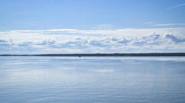 Berrak bir gökyüzü altında sakin bir deniz manzarası. Su hafif bulutları yansıtır İskandinav doğasını