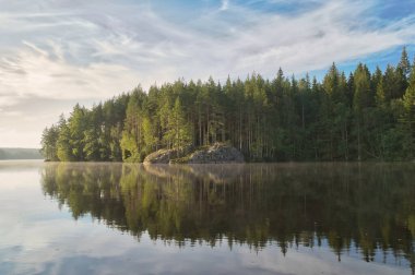 Yumuşak gün batımı ışığı sakin gölü yansıtır. Orman, kayalar ve gökyüzü barışçıl ve uyumlu bir manzara yaratmak için birleşir..