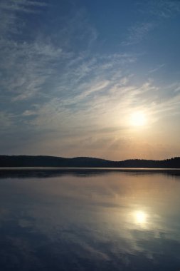 Sakin bir gölün üzerinde gün batımının atmosferik görüntüsü ve suda yansımalar. Yumuşak renkler ve huzurlu bir doğal atmosfer..