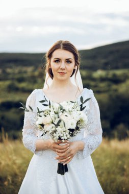 Doğanın ortasında beyaz elbiseli güzel bir gelinin fotoğrafı. Gelin elinde güzel bir buket tutuyor.