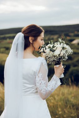 Doğanın ortasında beyaz elbiseli güzel bir gelinin fotoğrafı. Gelin elinde güzel bir buket tutuyor.