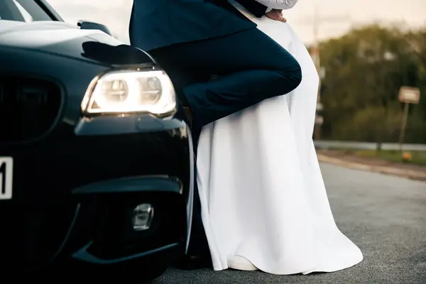 a man in a suit and a woman in a dress lean against a black car