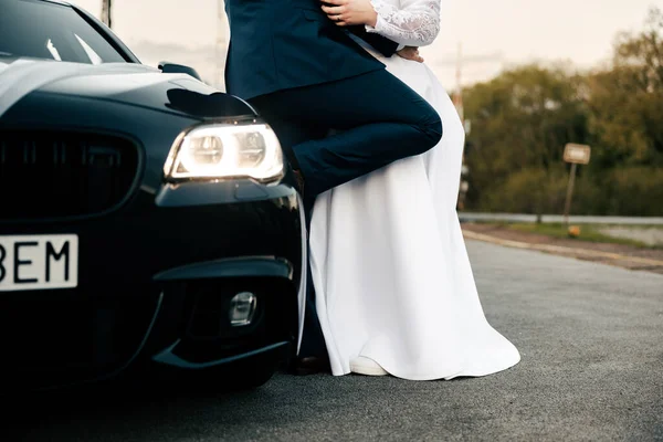 a man in a suit and a woman in a dress lean against a black car