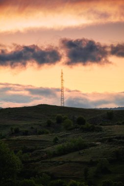 Tepenin tepesinde elektrik direği var. Arkasında parlak turuncu ışık var. Gün doğumunda..