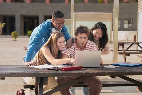 A group of four students with laptops and notes sit near the campus to review the exam.