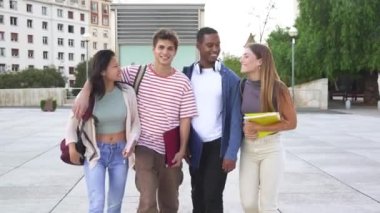 Çok ırklı bir grup öğrenci kameraya sarılıp yürüyorlar üniversitede açık havada gülüyorlar. Gençler birlikte eğleniyor..