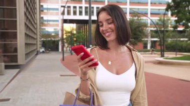 Genç bir kadın cep telefonunu kaldırıyor ve sokakta elinde alışveriş torbalarıyla konuşuyor..