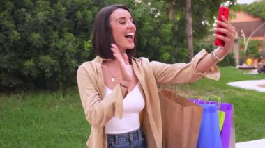 Alışveriş torbalarıyla sokakta oturan genç ve gülümseyen bir kadın video görüşmesi yapıyor.