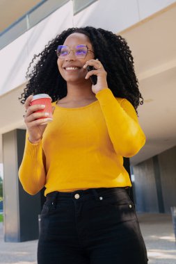 Dikey fotoğraf. Üniversite kampüsünde kahve içerken gülümseyen siyah öğrenci cep telefonuyla konuşuyor. Kavram: Üniversite hayatı