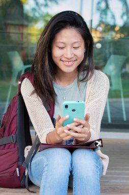 Güzel Çinli ergen öğrenci dışarıda cep telefonunda sosyal medya uygulaması kullanarak gülümsüyor. Cep telefonunda sohbet eden kız.
