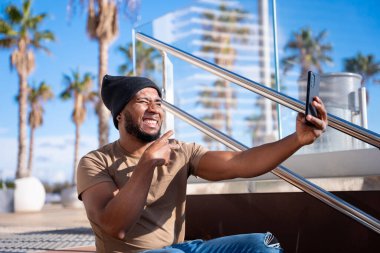 Mutlu siyah adam akıllı telefonuyla selfie çekiyor. Açık havada barış işareti yapan gülümseyen genç bir Latin 'in portresi..