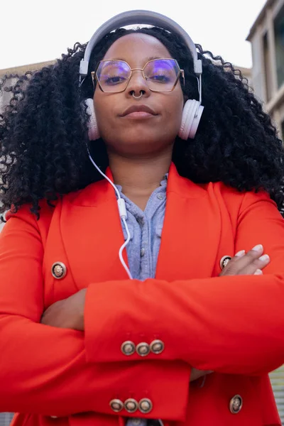 portrait black woman with headphones looking at a camera with arms crossed outdoors.