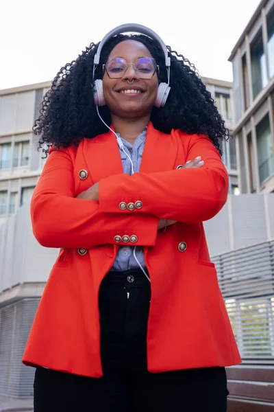 portrait happy black woman with headphones looking at a camera with arms crossed outdoors.