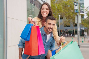 Mutlu beyaz adam kız arkadaşını sırtında taşıyor. Birlikte eğleniyor. Alışveriş torbaları taşıyor..