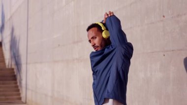  Spor kulaklık takan, esneme hareketleri yapan, açık havada kameraya gülümseyen genç bir kadın..
