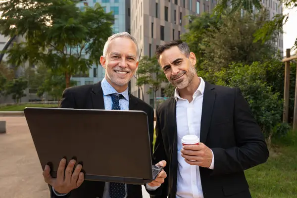 Portrait of two trusted business colleagues dressed in formal attire with a computer in hand laughing looking at camera.