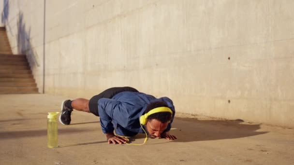 スポーツヘッドフォンで 床に水ボトルが付いているプッシュアップを行う集中した若い男 — ストック動画
