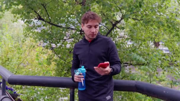 運動に疲れたアスリートの白人男性は 携帯電話アプリを使用している間 水を飲むために休憩を取ります アウトドア — ストック動画