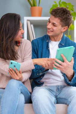  Genç, gülümseyen, çok kültürlü bir çift evdeki koltukta, bir cihazdaki kablosuz internet uygulamasını tarayan bir tablet ve cep telefonu kullanarak oturuyor. Teknoloji konsepti.