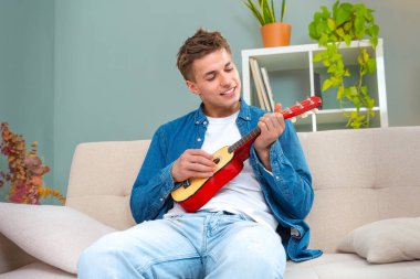 Genç adam evde ukulele çalıyor. Medya. Adam evde tek başına ukulele çalıyor. Genç adam kanepede otururken küçük bir gitar çalma pratiği yapıyor.