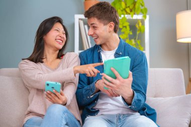  Genç, gülümseyen, çok kültürlü bir çift evdeki koltukta, bir cihazdaki kablosuz internet uygulamasını tarayan bir tablet ve cep telefonu kullanarak oturuyor. Teknoloji konsepti.