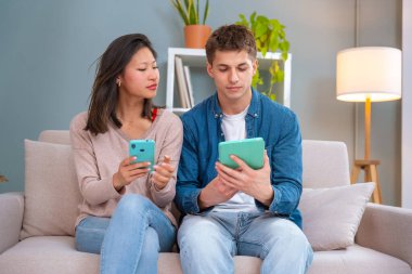  Genç, çok ırklı bir çift evdeki koltukta, bir cihazda kablosuz internet uygulamasını tarayan bir tablet ve cep telefonu kullanarak oturuyor. Teknoloji konsepti.