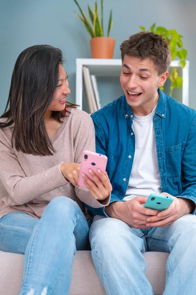  Genç, gülümseyen, çok kültürlü bir çift evdeki koltukta, bir cihazdaki kablosuz internet uygulamasını tarayan bir tablet ve cep telefonu kullanarak oturuyor. Teknoloji konsepti.