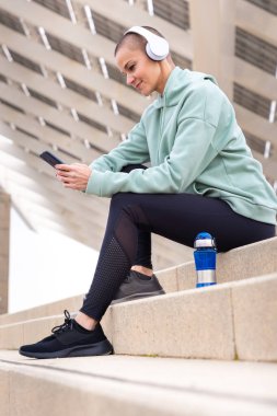 Egzersiz yapmaktan yorgun kulaklıklı atletik bir kadın cep telefonu uygulaması kullanarak su molası vermek için oturur..