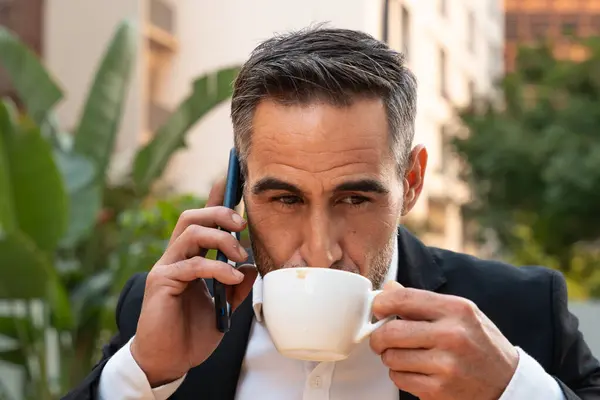 Middle aged man sitting drinking coffee happily having a work conversation with cell phone.