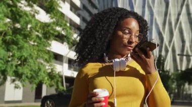 Gözlüklü ve kulaklıklı mutlu genç siyahi kadın, elinde kahveyle cep telefonuyla elsiz sohbet ediyor..