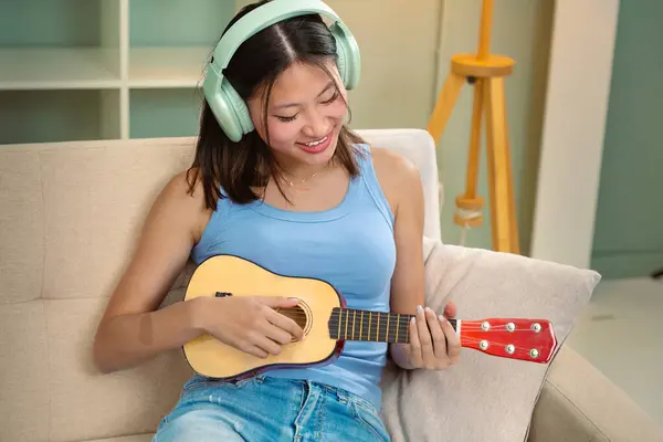 The young woman plays the ukulele at home. Media. The woman plays the ukulele alone at home. Young woman practices playing a small guitar while sitting on the couch