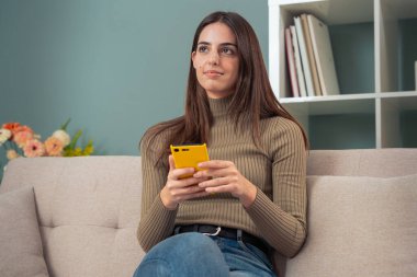 Kanepede güzel, düşünceli bir kadın, kolay ve güvenli elektronik ödemeler için internetten cep telefonuyla mutlu alışverişler. İçeride.