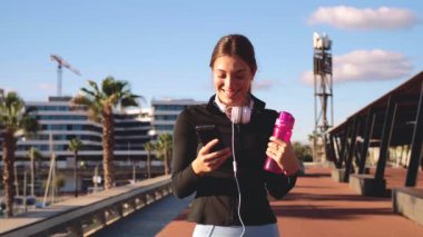 Neşeli genç bir kadın elinde bir spor şişesiyle cep telefonuyla sohbet ediyor.