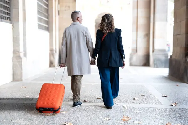 Gülümseyen orta yaşlı beyaz turist çifti bavullarıyla el ele yürürken birbirlerine bakıyorlar. Mutlu karı koca güneşli bir kış gününde dışarıda tatilin tadını çıkarıyorlar.