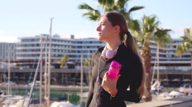 Neşeli genç bir kadın bir arkadaşıyla konuşmak için akıllı telefonu kaldırıyor, diğer elinde bir spor şişesi tutuyor..