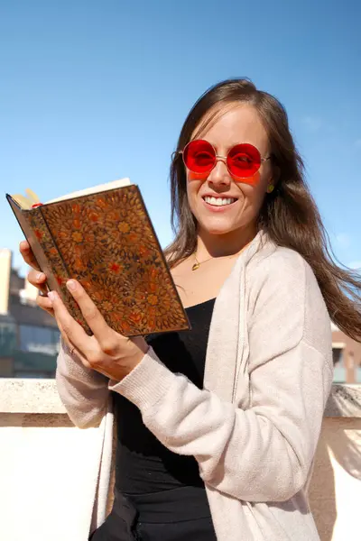 Yoğunlaşmış beyaz bir kadın kameraya bakarak kitap okuyor..