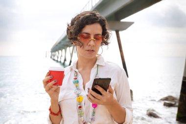 Kafkasyalı bir kadının deniz kenarında tatil yaparken güneş gözlüğü takıp cep telefonuyla sosyal medya uygulaması kullanarak gülerken görüntüsü.. 