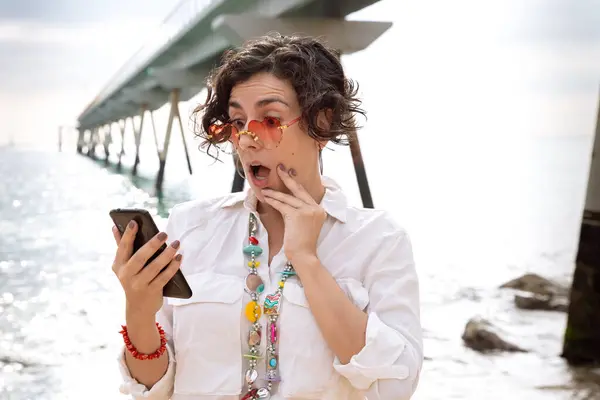 Deniz kenarında tatil yapan, güneş gözlüğü takan beyaz bir kadın cep telefonuyla sosyal medya uygulaması kullanarak sürpriz bir hareket yaptı.