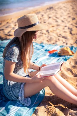  Sahilde oturan, şapkalı, arka planda deniz olan bir kadın kitap okuyor.