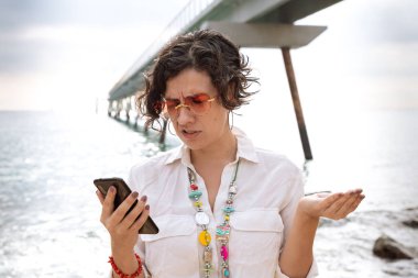 Caucasian woman wearing sunglasses vacationing by the sea with a gesture of surprise using a social media app with her mobile phone clipart