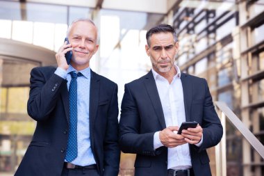 İş görüşmesi sırasında iş yerinin dışında birlikte yürürken cep telefonuyla konuşan ve uygulama kullanan iki güvenilir iş arkadaşı.