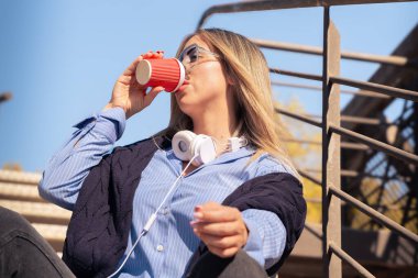 Üniversite kampüsünde kahve içerken gülümseyen kulaklı ve gözlüklü genç bir öğrenci. Konsept: Üniversite hayatı