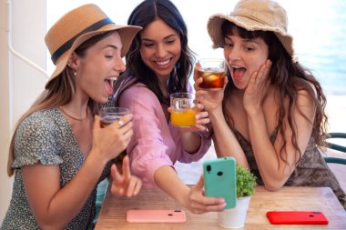 Bir grup gülen etnik kadın bir restoranda tatilin tadını çıkarıyor. Güzel ve neşeli Z Nesli kızları internette akıllı telefonlarıyla geziniyor.