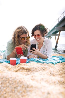 Sahilde oturan orta yaşlı bir adam ve kadın şaşırmış bir suratla akıllı telefon uygulamalarına göz atıyorlar. Kavram: Tatil ve Teknoloji