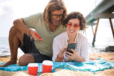 Sahilde oturan orta yaşlı bir adam ve kadın şaşırmış bir suratla akıllı telefon uygulamalarına göz atıyorlar. Kavram: Tatil ve Teknoloji