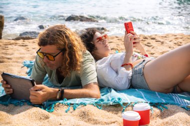 Sahilde oturan orta yaşlı bir adam ve kadın şaşırmış bir suratla akıllı telefon uygulamalarına göz atıyorlar. Kamera Konsepti: Tatil ve Teknoloji